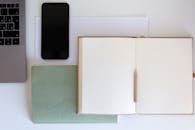 极简主义工作空间的顶视图中，白色办公桌上摆放着打开的笔记本、铅笔、笔记本电脑和智能手机