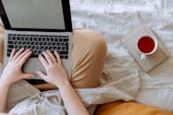 顶视图中，一位匿名的远程工作人员盘腿坐在床上，一边在家喝茶，一边在笔记本电脑的空白屏幕上打字