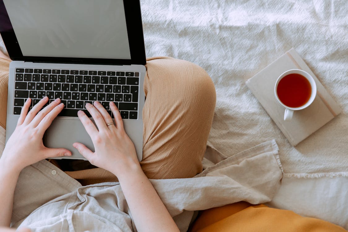 Free Top view anonymous remote worker typing on keyboard of laptop with blank screen while sitting with crossed legs on bed with tea aside at home Stock Photo