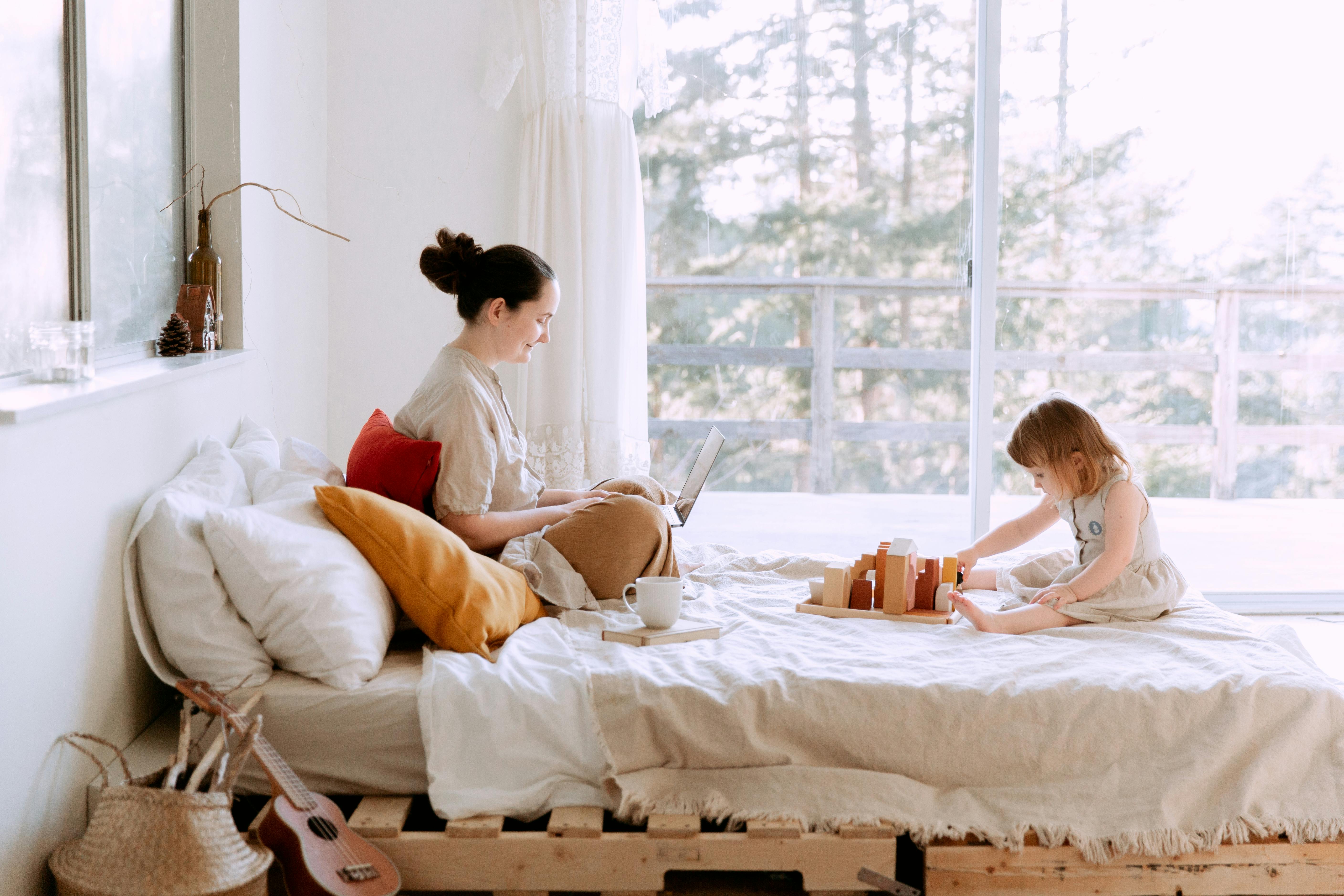 full body of happy mother surfing netbook and daughter playing with toys on bed against window