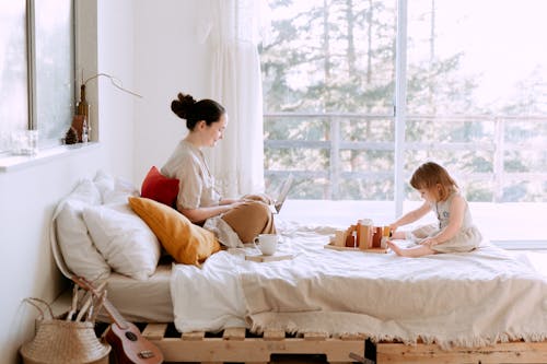 Free Full body of happy mother surfing netbook and daughter playing with toys on bed against window Stock Photo