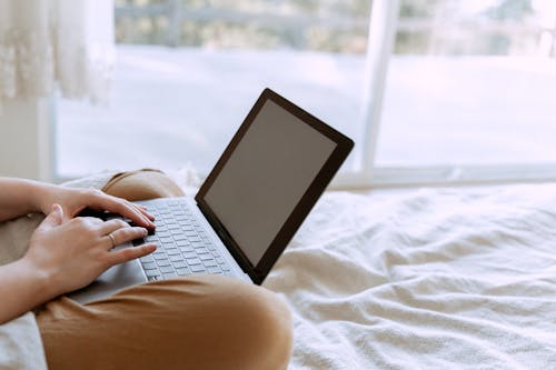 Free Crop faceless person in casual wear using netbook with empty screen while chilling on bed in lotus pose near window at home Stock Photo