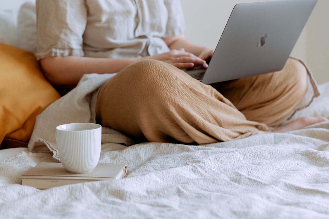 Crop woman lounging with laptop and cup of coffee on bed at home