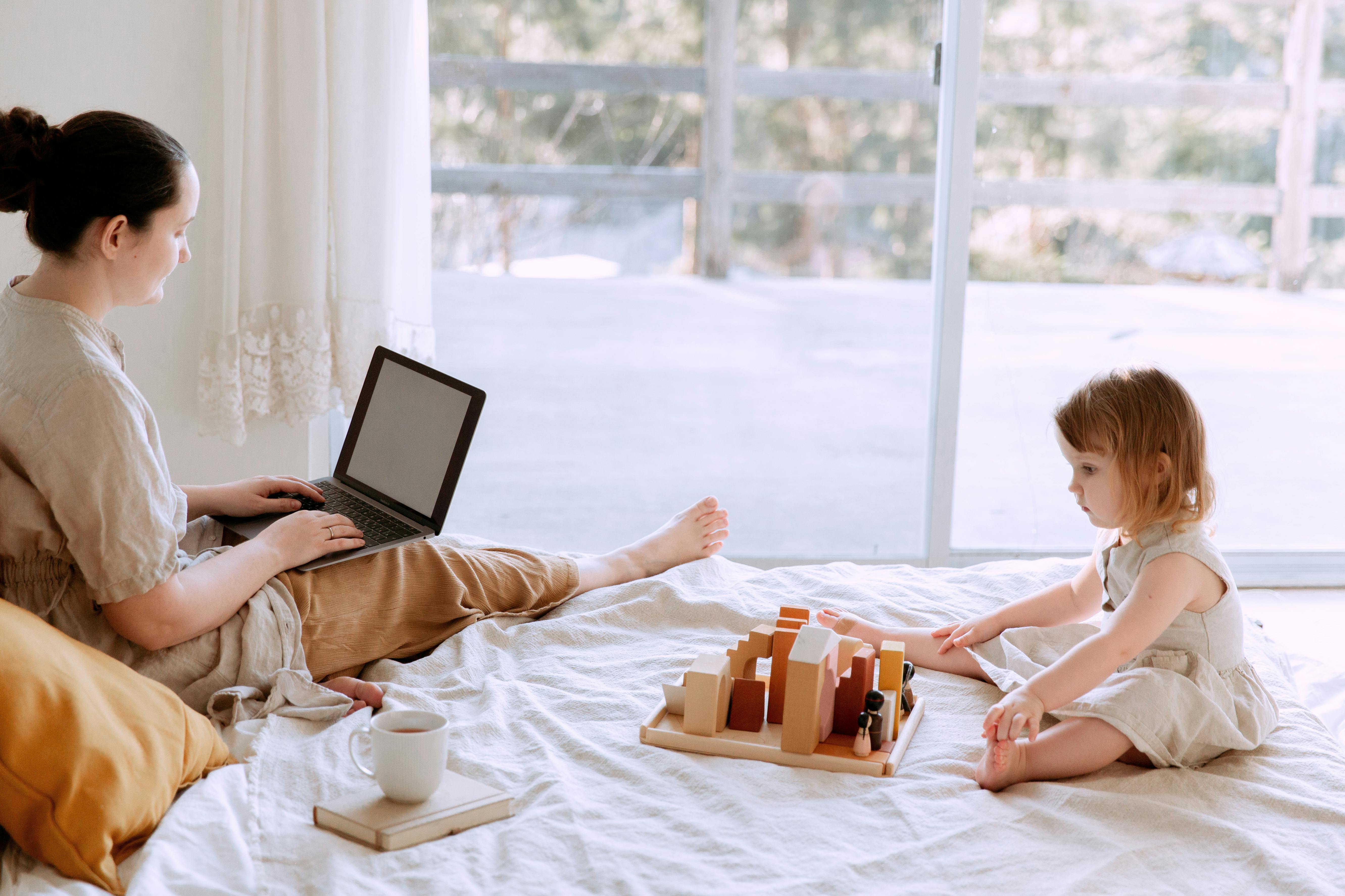 mother using laptop while cute little girl playing on bed at home