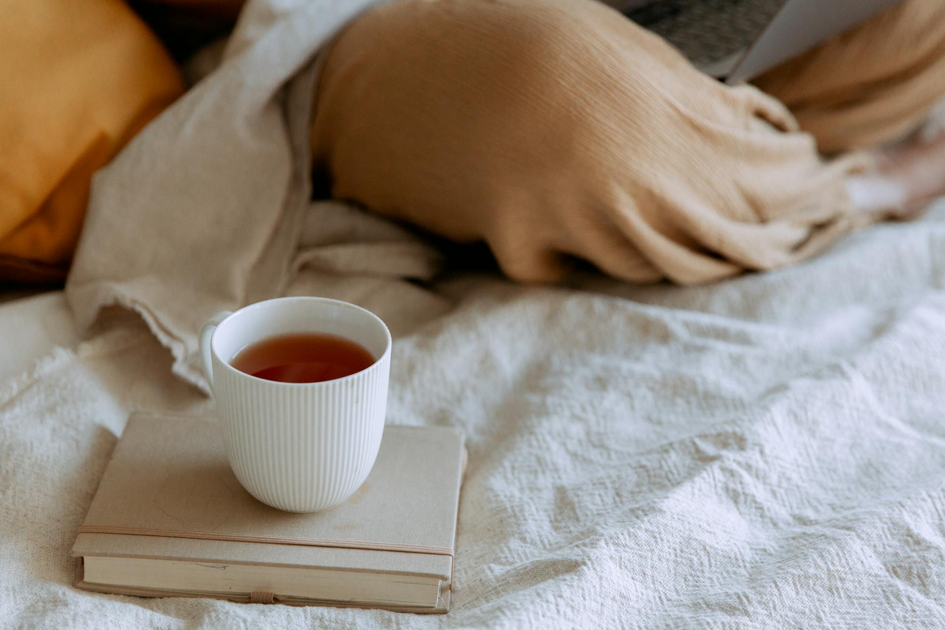 From above of tea placed on notepad near freelancer sitting with crossed legs using netbook on bed at home