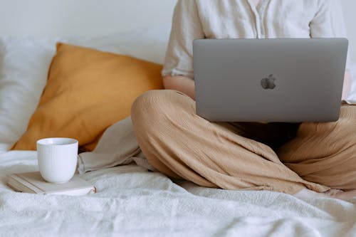 Free Crop faceless female in casual outfit sitting on bed with legs crossed holding laptop on knees with cup of coffee standing on notebook while working from home Stock Photo