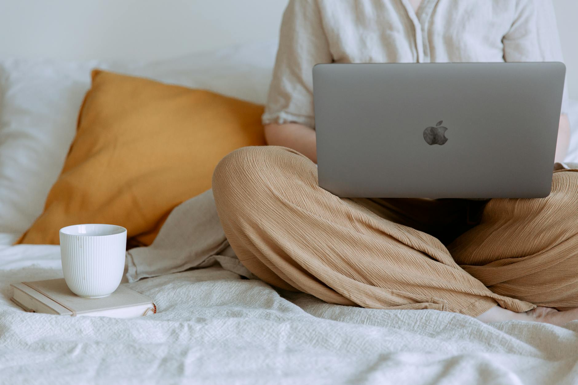 Crop faceless female in casual outfit sitting on bed with laptop