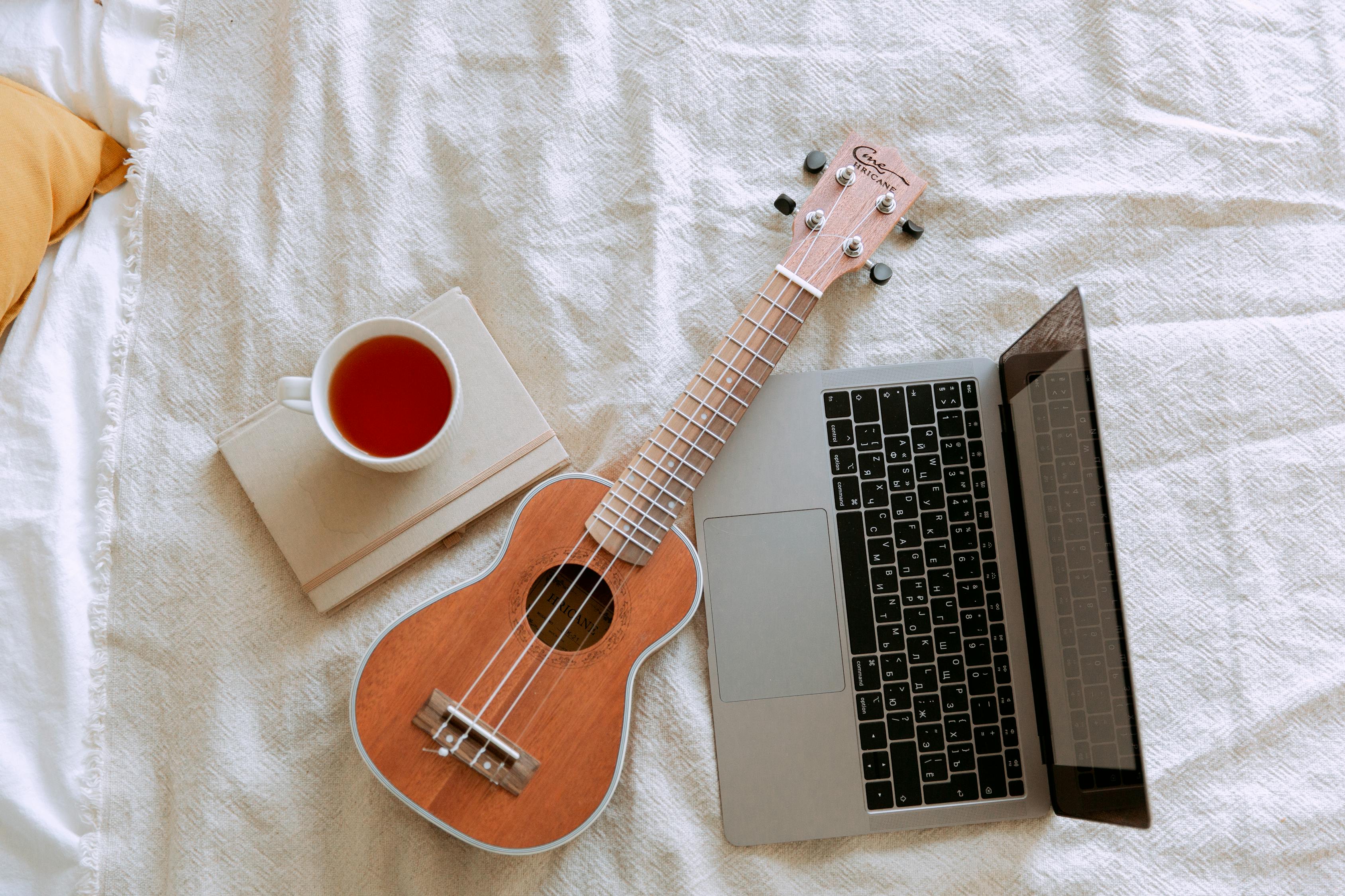A laptop resting on a bedsheet with a cup of tea and a ukulele