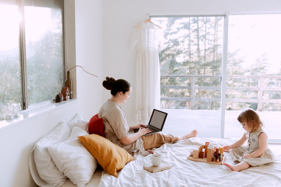 Side view of cute toddler girl sitting on bed barefoot and playing with colorful wooden blocks while mother using laptop in bed enjoying morning coffee