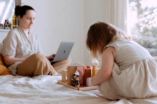 Free Side view of adorable toddler girl sitting on bed and playing with wooden toys while mother working from home with laptop on knees on sunny morning Stock Photo