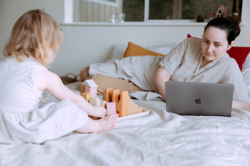 Free Cute toddler girl playing with wooden blocks on bed while mother using laptop nearby Stock Photo