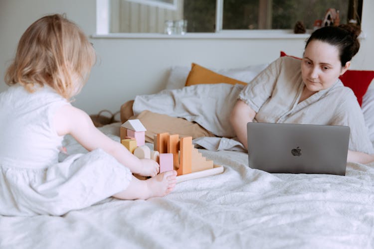 Cute Toddler Girl Playing With Wooden Blocks On Bed While Mother Using Laptop Nearby