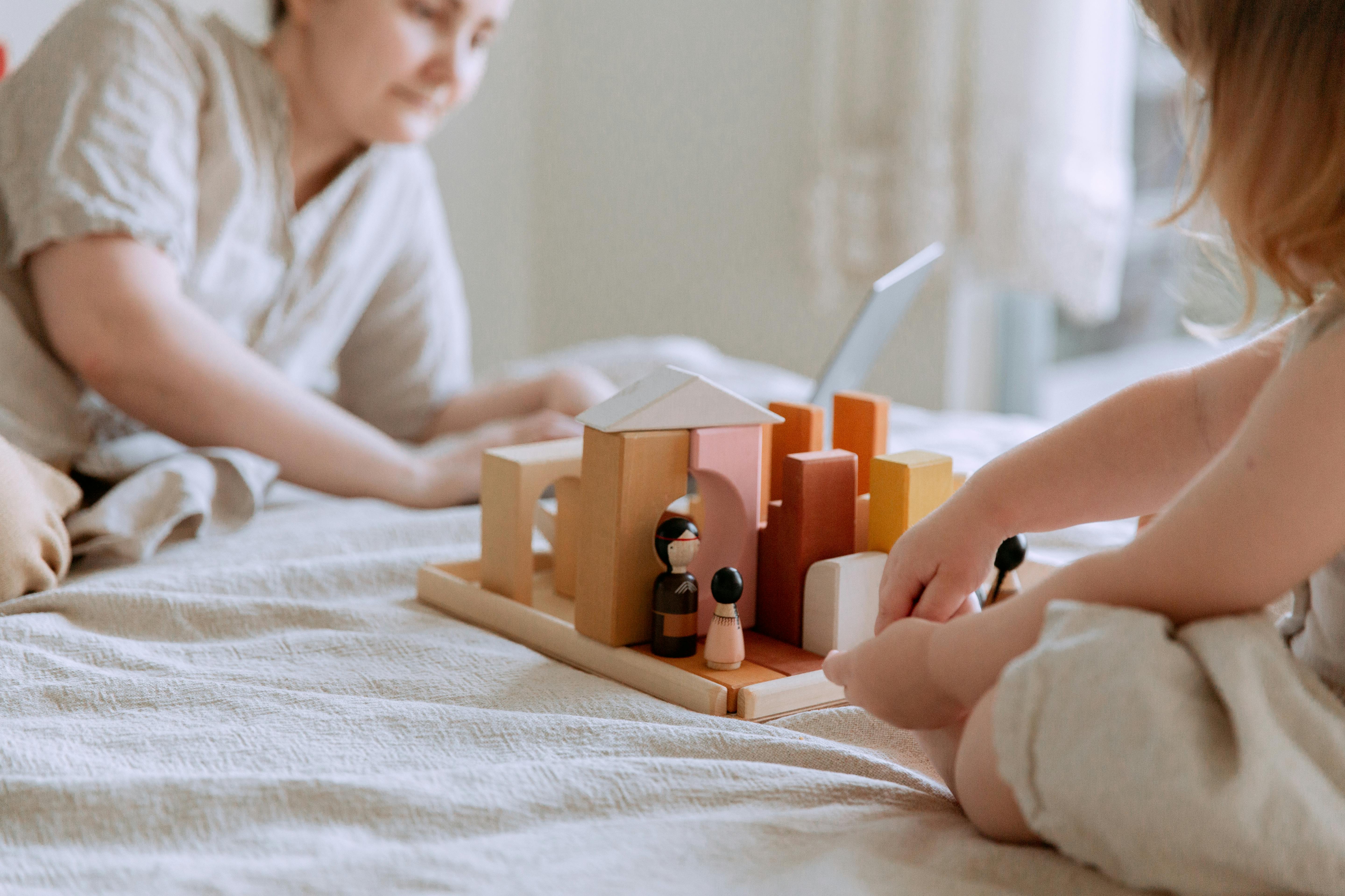 faceless toddler girl sitting on bed and playing with wooden blocks and toys while mother using laptop