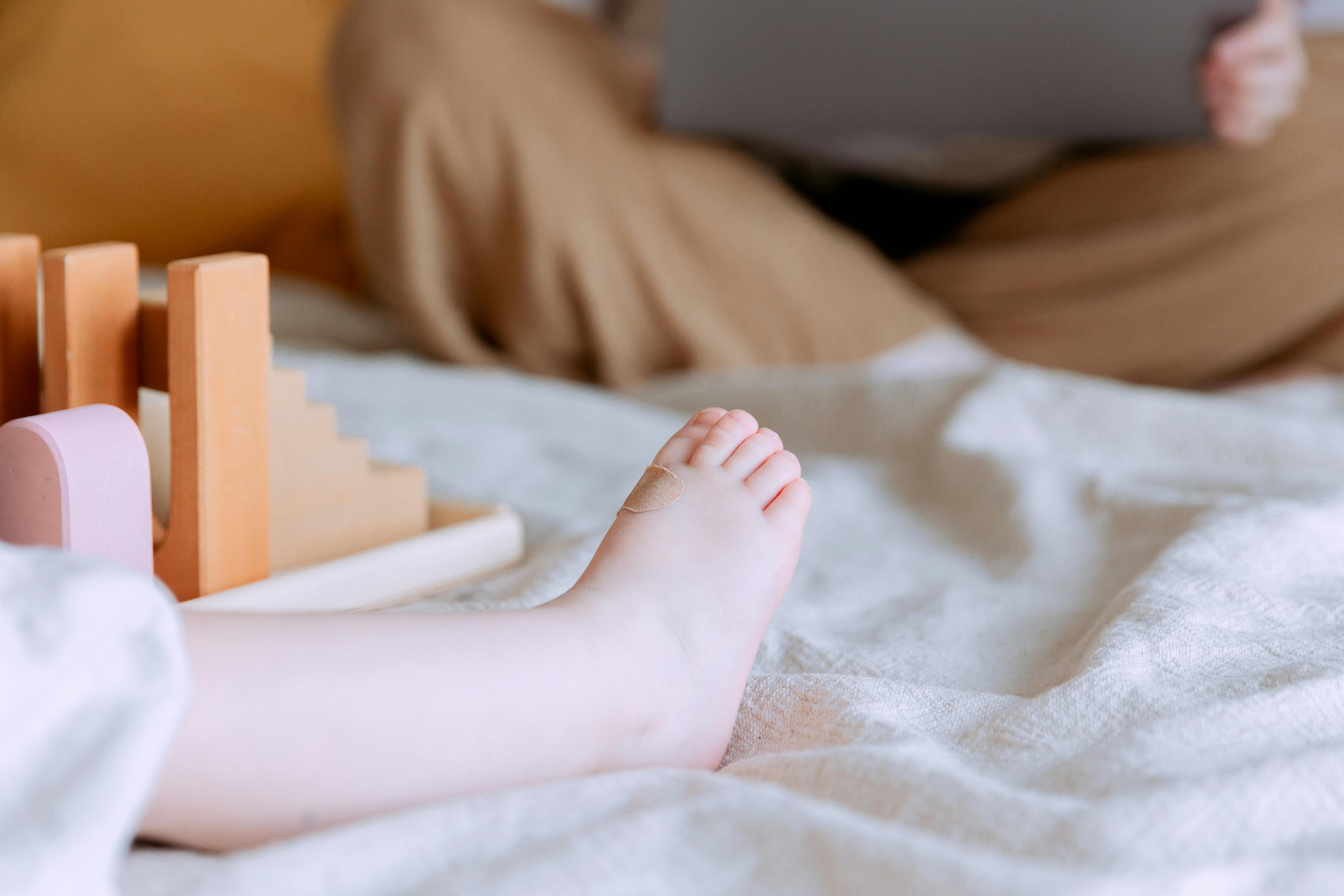 unrecognizable toddler sitting on bed with wooden constructor while mother using laptop