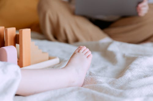 Crop faceless toddler sitting on bed with colorful wooden building blocks while mother sitting with legs crossed doing remote work with laptop on knees on background