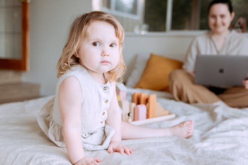 Cute toddler girl sitting and playing on bed while mother using laptop on background