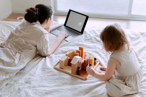 Free From above of young female in casual clothes lying on bed and using laptop while daughter sitting on bed and playing with wooden blocks and toys on sunny morning Stock Photo