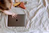 From above of crop faceless child sitting on bed with scattered multicolored details of wooden constructor and touching closed laptop with hand