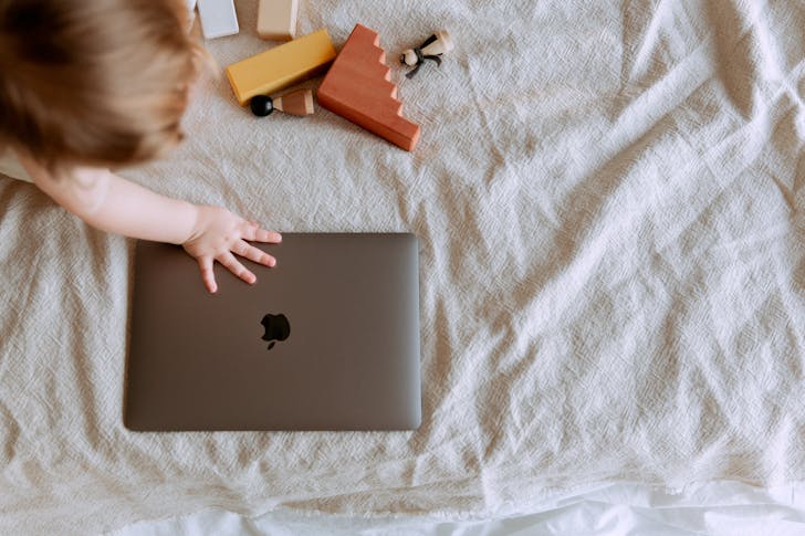 From above of crop faceless child sitting on bed with scattered multicolored details of wooden constructor and touching closed laptop with hand