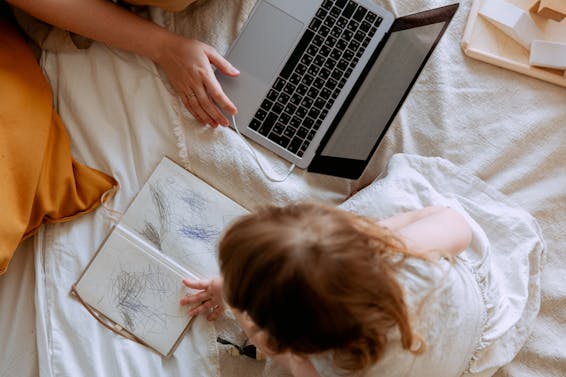 Top view of crop female using laptop while little girl drawing in notebook both chilling on cozy bed at home