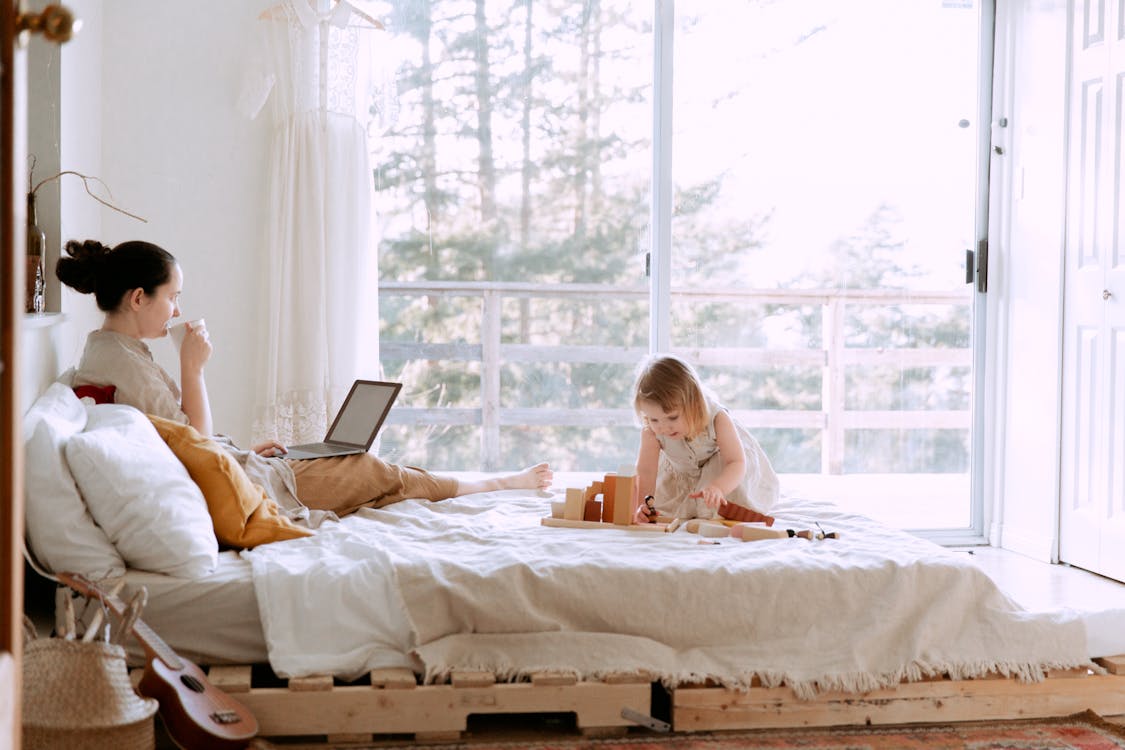 Concentrated female drinking coffee while working on project on laptop and daughter sitting on bed playing with constructor in bedroom