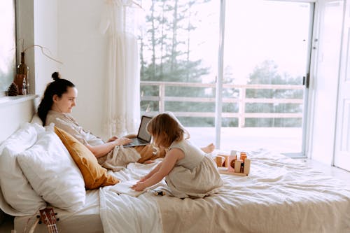 Free Side view of adult female and little girl wearing casual clothes sitting on bed and browsing laptop and drawing in notepad Stock Photo