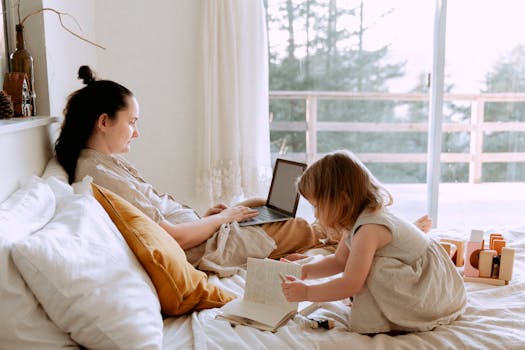A mother works remotely on a laptop while her child plays next to her in a cozy bedroom setting.