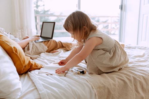 Free Side view of charming girl drawing in notebook while sitting on bed with crop mother freelancer using laptop Stock Photo