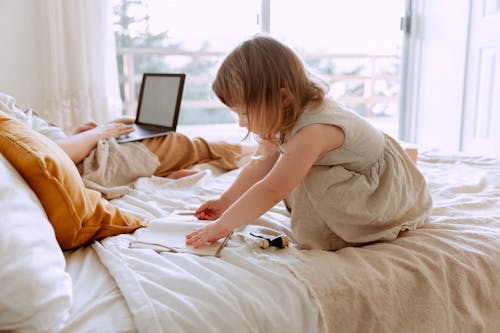 Side view of charming girl drawing in notebook while sitting on bed with crop mother freelancer using laptop