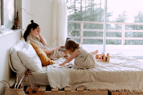 Side view of woman working with laptop remotely and spending time with daughter while resting on bed in cozy bedroom