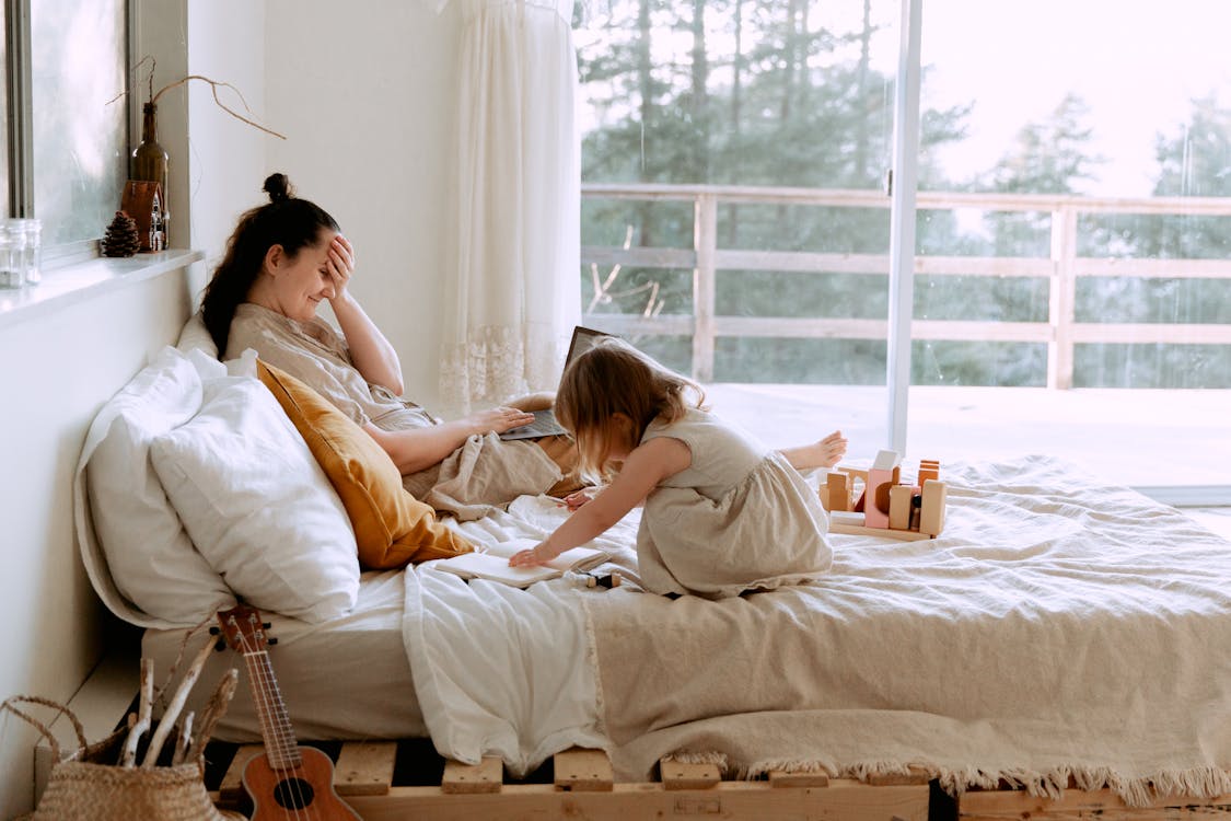 Side view of woman working with laptop remotely and spending time with daughter while resting on bed in cozy bedroom