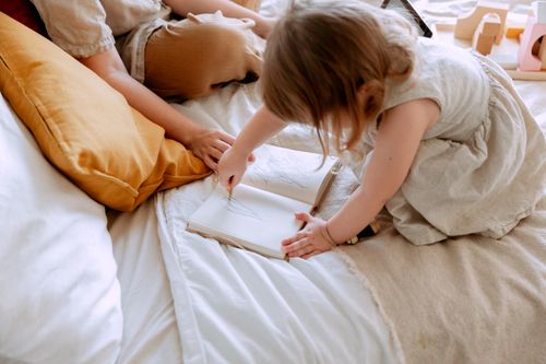 Free From above of small girl in dress drawing sketches in notebook with mother sitting near on bed Stock Photo