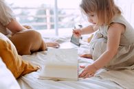 Daughter in dress drawing in notebook while mother working on laptop in bedroom