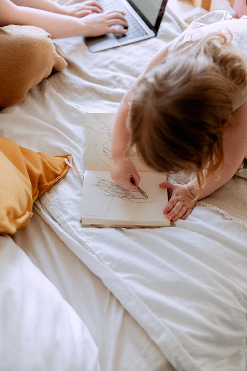 Free From above of small girl in dress drawing with pencil in notebook while mother using laptop on bed near Stock Photo