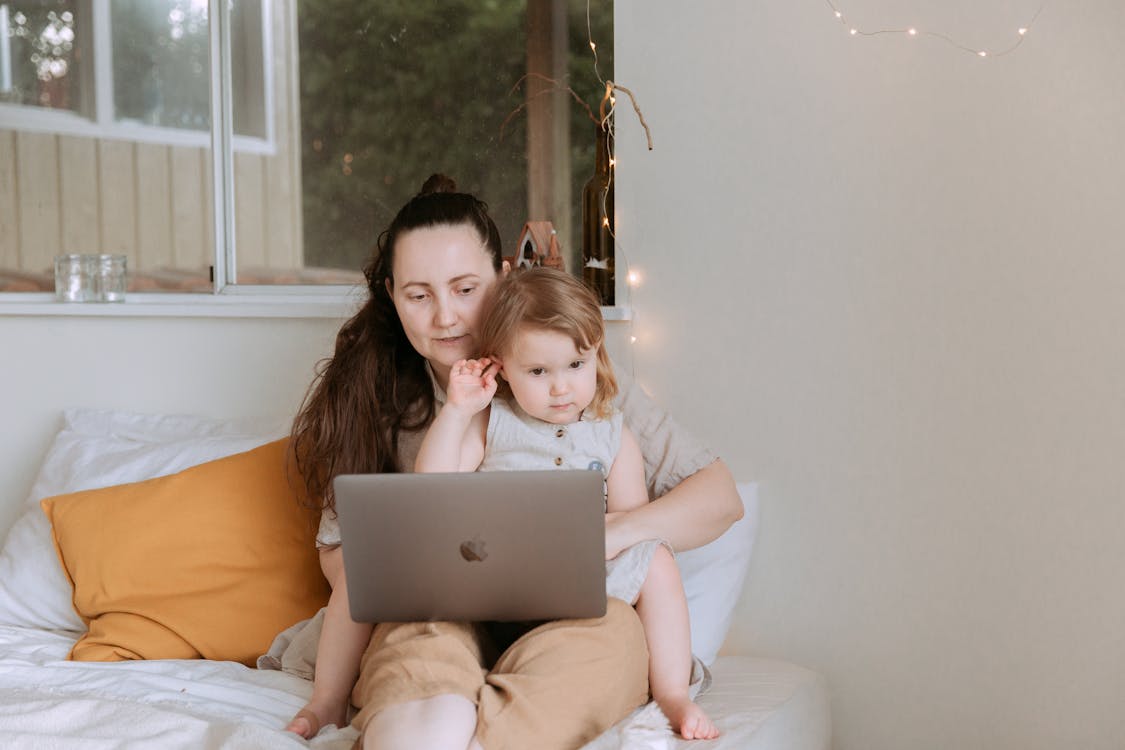 Adult woman with preschool girl in dress spending time together on bed using laptop