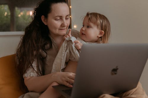Free Busy mother using laptop while holding in hand restless small daughter while sitting on sofa Stock Photo