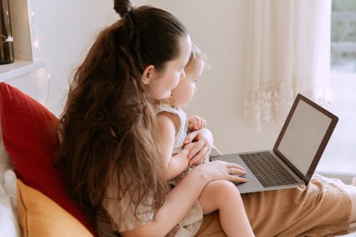 Free Side view of smiling mother cuddling little daughter while sitting on bed and typing on portable computer and looking at empty screen in afternoon at home Stock Photo