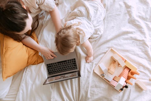 Top view of anonymous mother and little daughter in casual wear searching pictures on netbook while sitting on bed near educational toys in flat