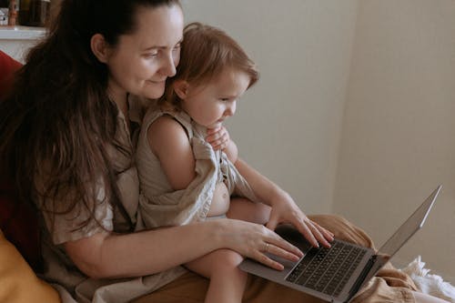 Free Crop side view of adult smiling mother embracing little daughter while typing on netbook and sitting in room in flat Stock Photo