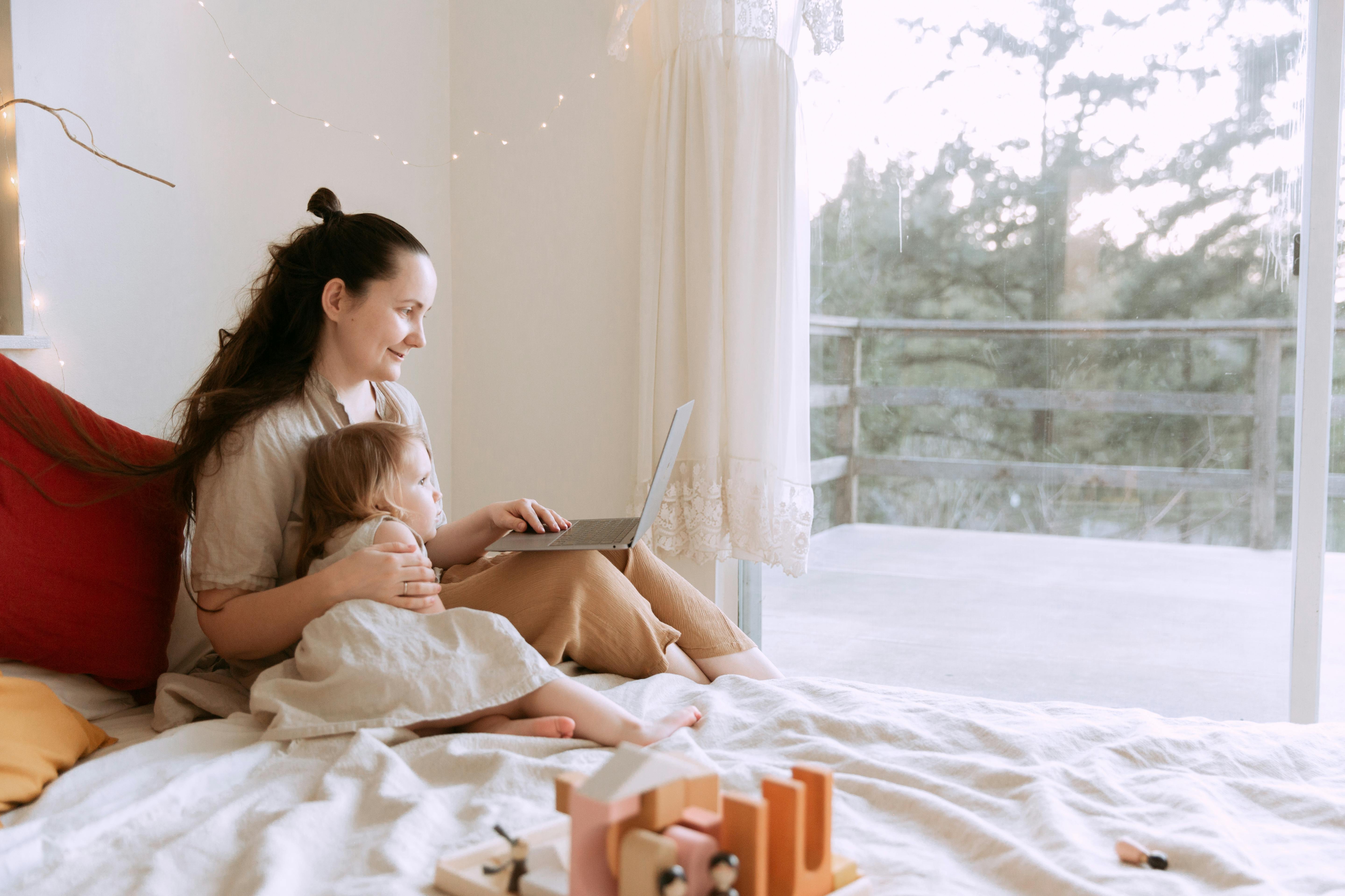 cheerful mother cuddling daughter while sitting on bed with laptop