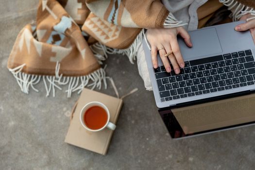 From above of crop anonymous remote worker in shawl with pencil using portable computer while sitting on concrete floor near cup of tea