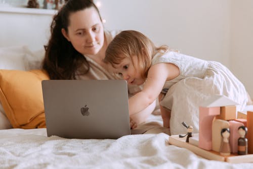 Adult mother with content little daughter sitting on comfortable bed in apartment near educational toys while surfing internet on portable computer at home