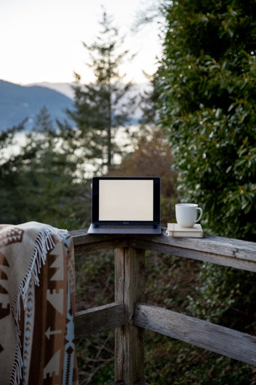 Free Portable computer with blank screen put on wooden fence near cup of hot drink and warm shawl behind mountains and trees in afternoon in countryside Stock Photo