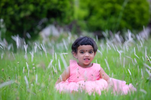 Free Charming child in a pink dress seated in lush green grass on a sunny day. Stock Photo