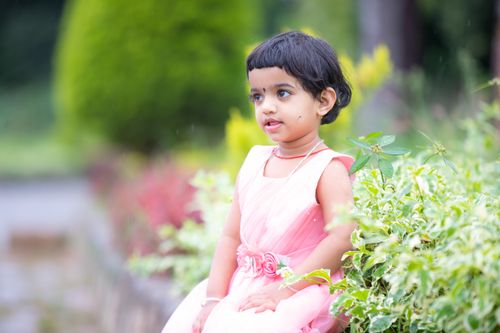 Free Adorable toddler sitting amidst lush greenery wearing a pink dress, captured in a serene garden setting. Stock Photo