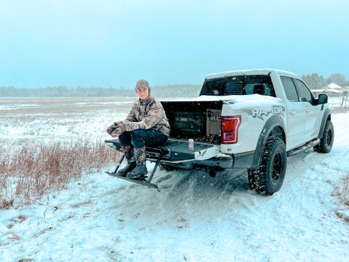 Free Person sitting on a truck tailgate in a snowy field, dressed in winter camo gear. Stock Photo