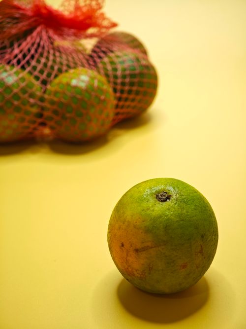 Free Orange fruit with a vibrant yellow backdrop, featuring one orange in focus and a netted bag in soft focus. Stock Photo