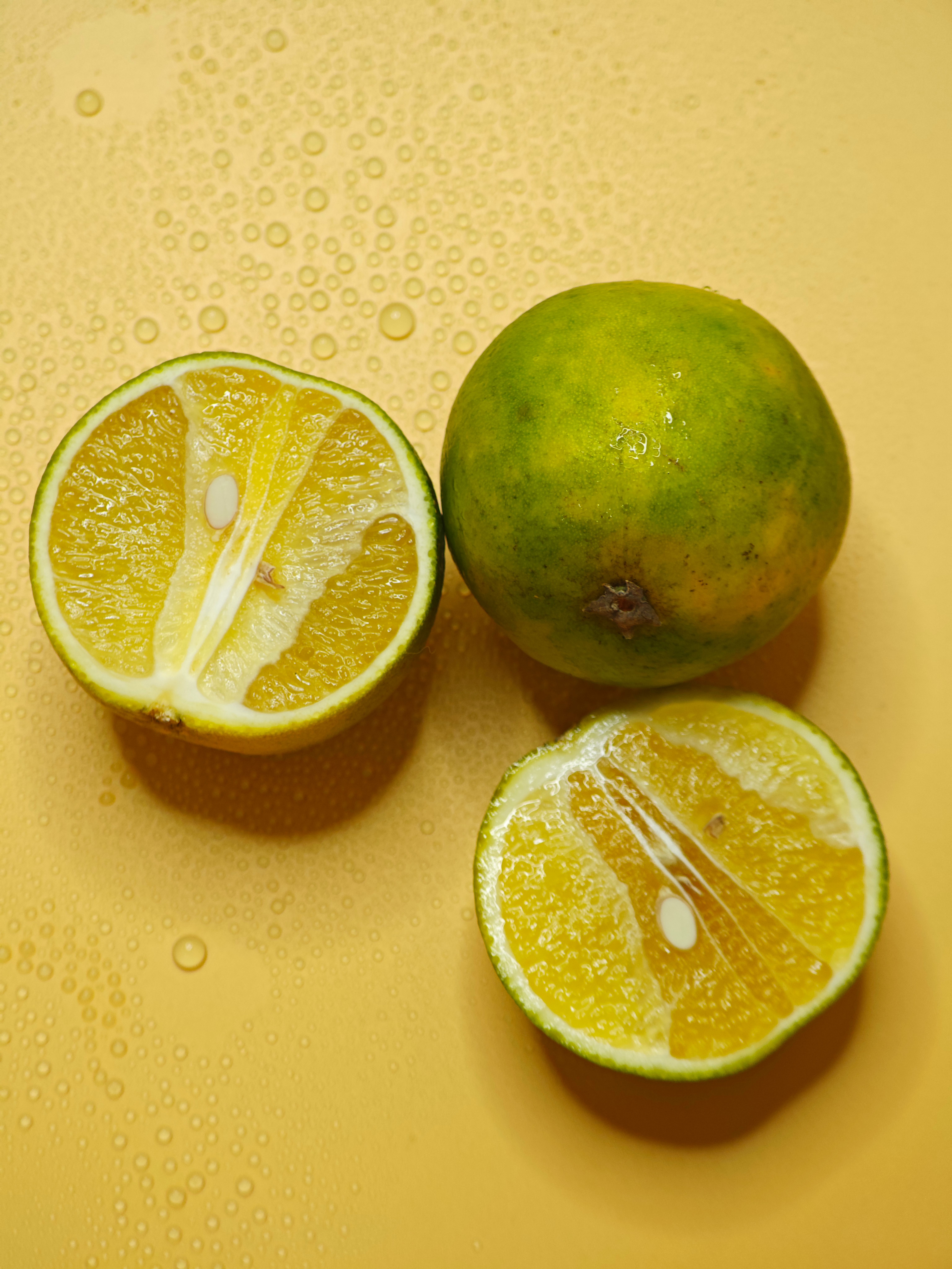 Whole and halved citrus fruits on a yellow surface with water droplets.
