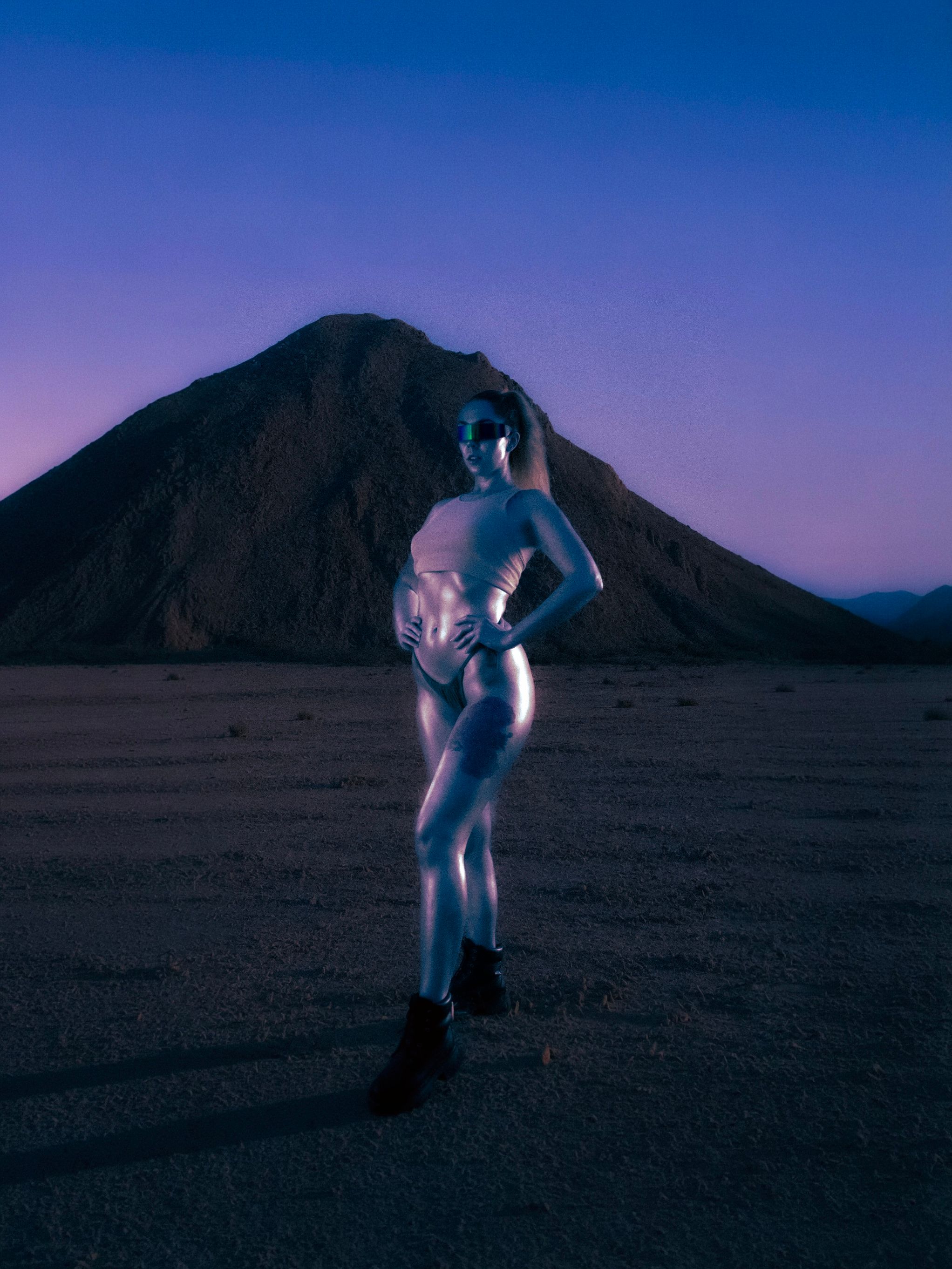 A futuristic model poses in metallic attire against a desert backdrop at twilight, exuding a sci-fi vibe.