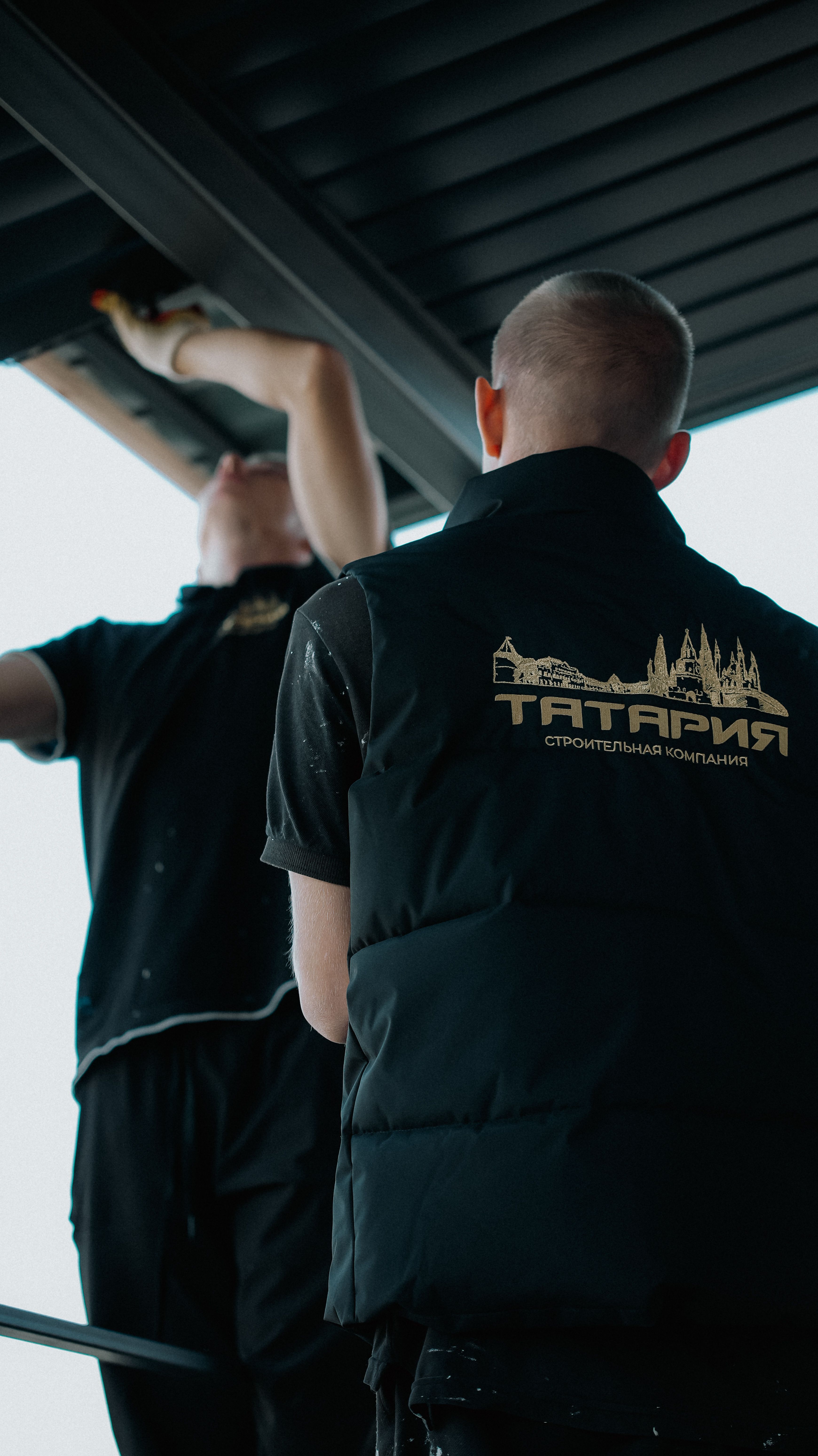 Two construction workers install roof panels using tools, highlighting teamwork and craftsmanship.